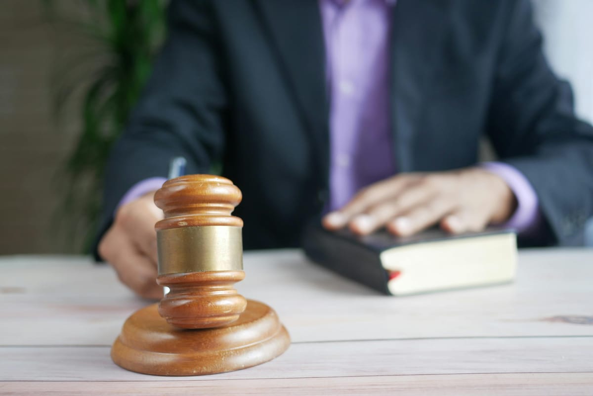 Close-up of a judge’s gavel on a desk, with a person in formal attire and a law book blurred in the background.
