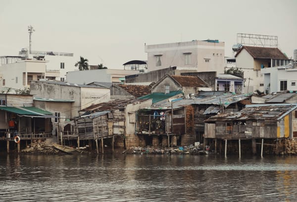 A waterfront settlement with weathered wooden homes on stilts in the foreground and larger concrete buildings behind them und